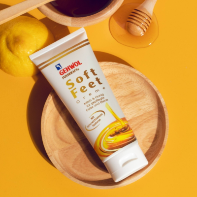 Tubo de creme GEHWOL Soft Feet numa prato de madeira com limão e colher de mel ao lado, sobre fundo amarelo