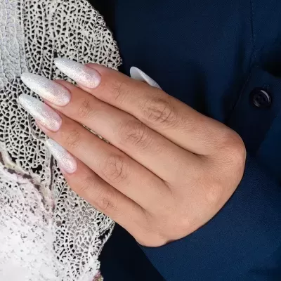 Unhas artificiais prateadas e brilhantes em mão com fundo escuro e tecido branco rendado