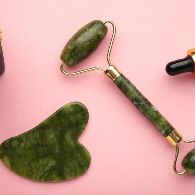 Rolo de massagem facial verde, gua sha verde e conta-gotas com tampa dourada sobre fundo rosa
