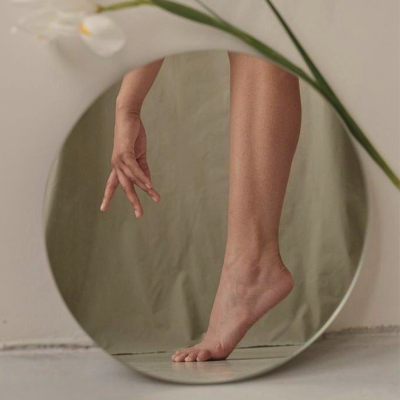 Reflexo de perna e pé descalço feminino em espelho redondo com moldura branca e flor branca.