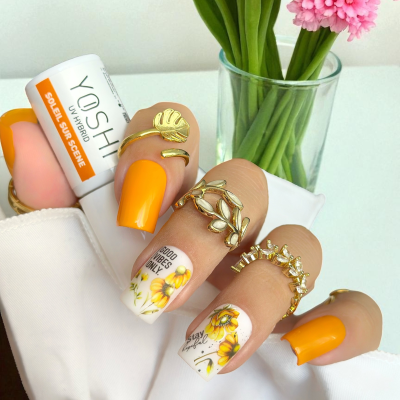 Unhas decoradas com esmalte laranja e branco com flores amarelas, anéis dourados e protetor solar Yoshi ao lado.