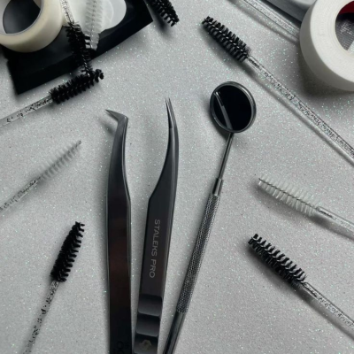 Ferramentas para estética com pinças pretas, espelho e escovas de sobrancelhas sobre fundo branco.