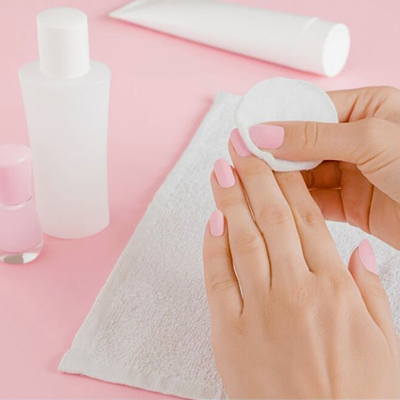 Mãos com unhas pintadas rosa claro segurando disco de algodão sobre toalha branca com frascos translúcidos em fundo rosa