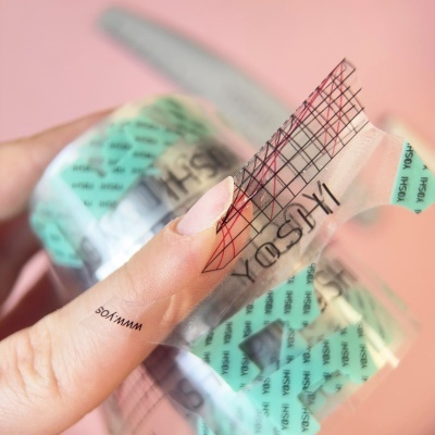 Fita adesiva transparente com padrão e texto YOSHI em fundo branco, segurada por mão com unhas naturais