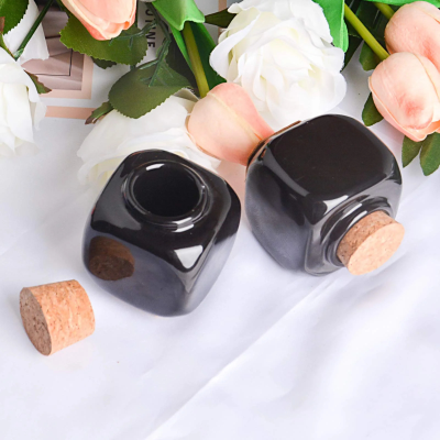 Dois frascos quadrados pretos com tampas de cortiça em fundo branco com flores artificiais.