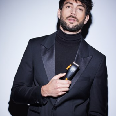 Homem com casaco preto e camisola preta segurando máquina de cortar cabelo preta com detalhes amarelos