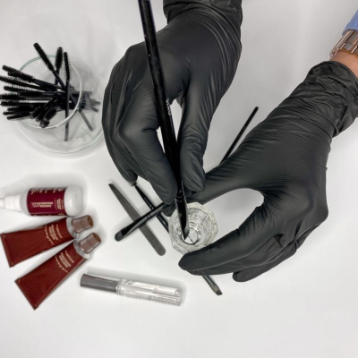 mãos com luvas pretas segurando pincel e vidro transparente com líquido, tubos vermelhos e aplicadores pretos ao lado