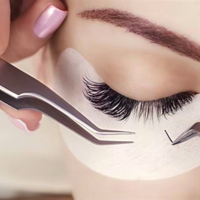 Mão feminina segurando pinça prateada aplicando pestana postiça negra sobre almofada branca no olho fechado