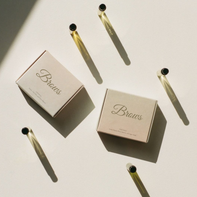 Duas caixas de papel bege com texto 'Brows' em dourado e cinco frascos pequenos com tampas pretas sobre superfície branca
