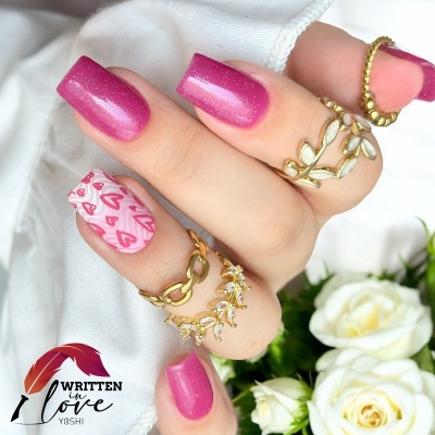 Mão feminina com unhas cor de rosa e padrão de corações, anéis dourados e flores brancas ao fundo
