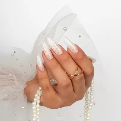 Unhas artifciais translúcidas com brilho, mão com anel dourado e colar de pérolas, fundo branco com tecido decorado com pérolas