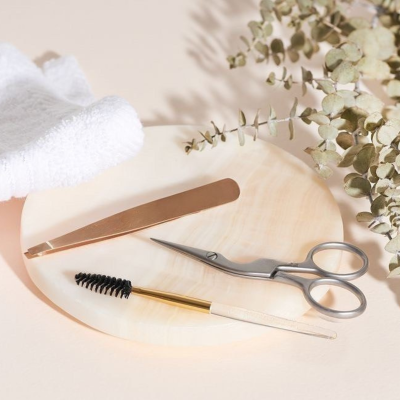 Conjunto de cuidados para sobrancelhas com pinça, escova e tesoura em prato claro.