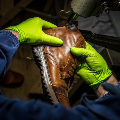 Sapato de couro castanho com sola branca e preta sendo costurado com luvas verdes