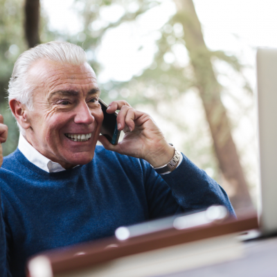 Homem mais velho em camisola azul e camisa branca a falar ao telemóvel junto a computador portátil cinzento numa mesa ao ar livre
