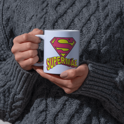 Caneca branca com texto SUPER MÃE com logo tipo super-herói em fundo suéter cinza