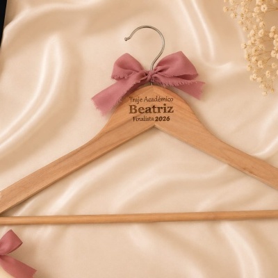Cabide de madeira personalizado com laço rosa e texto para traje académico, com diploma e faixa de finalista, sobre tecido creme.