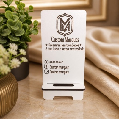 Suporte para telemóvel branco com logo e texto da Custom Marques numa mesa de mármore com plantas de decoração ao lado.