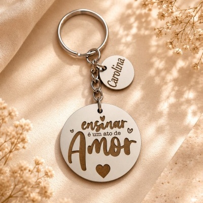 Chaveiro redondo metal prateado com gravação ensinar é um ato de Amor e pendente menor com nome Carolina em fundo bege com flores secas.
