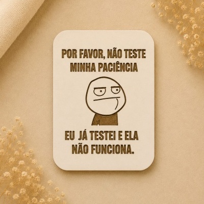 Placa decorativa de madeira com texto gravado e desenho de meme sobre fundo bege com flores secas.