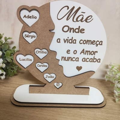 Objeto decorativo em madeira e branco com formato de rosto e corações com nomes e frase para mãe