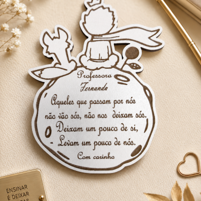 Placa decorativa em madeira branca com ilustração do Pequeno Príncipe e raposa e textos em português, sobre superfície bege com decoração seca e objetos dourados.