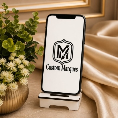 Smartphone preto num suporte branco junto a vaso com planta e flores sobre mesa de mármore.