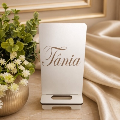 Suporte de mesa branco com nome Tânia em dourado junto a vaso com plantas e tecido dourado