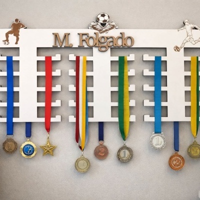 Suporte branco para medalhas com figuras de futebol e nome M. Folgado, com medalhas coloridas penduradas.