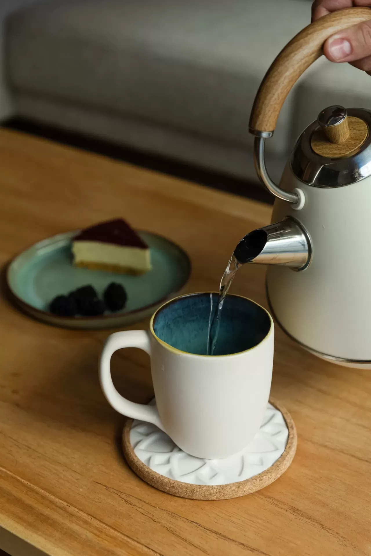Bule branco com pega de madeira e caneca branca a receber água sobre mesa de madeira com prato ao fundo.