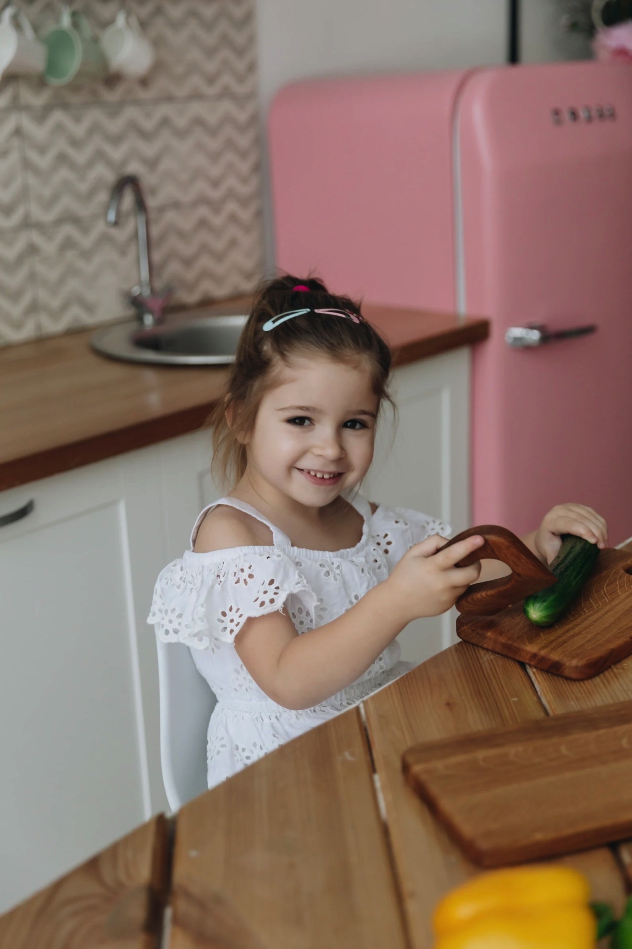Criança com vestido branco a cortar pepino em cozinha com frigorífico rosa