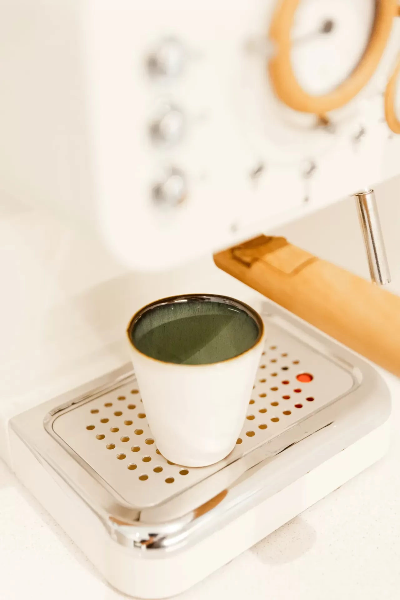 Máquina de café branca com detalhes em madeira clara e um copo cerâmico com interior verde.