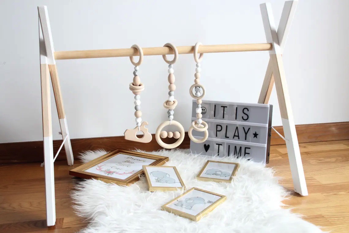 Estrutura de madeira para brinquedos de bebé com três brinquedos pendurados, quadro com texto IT IS PLAY TIME e quatro molduras no chão