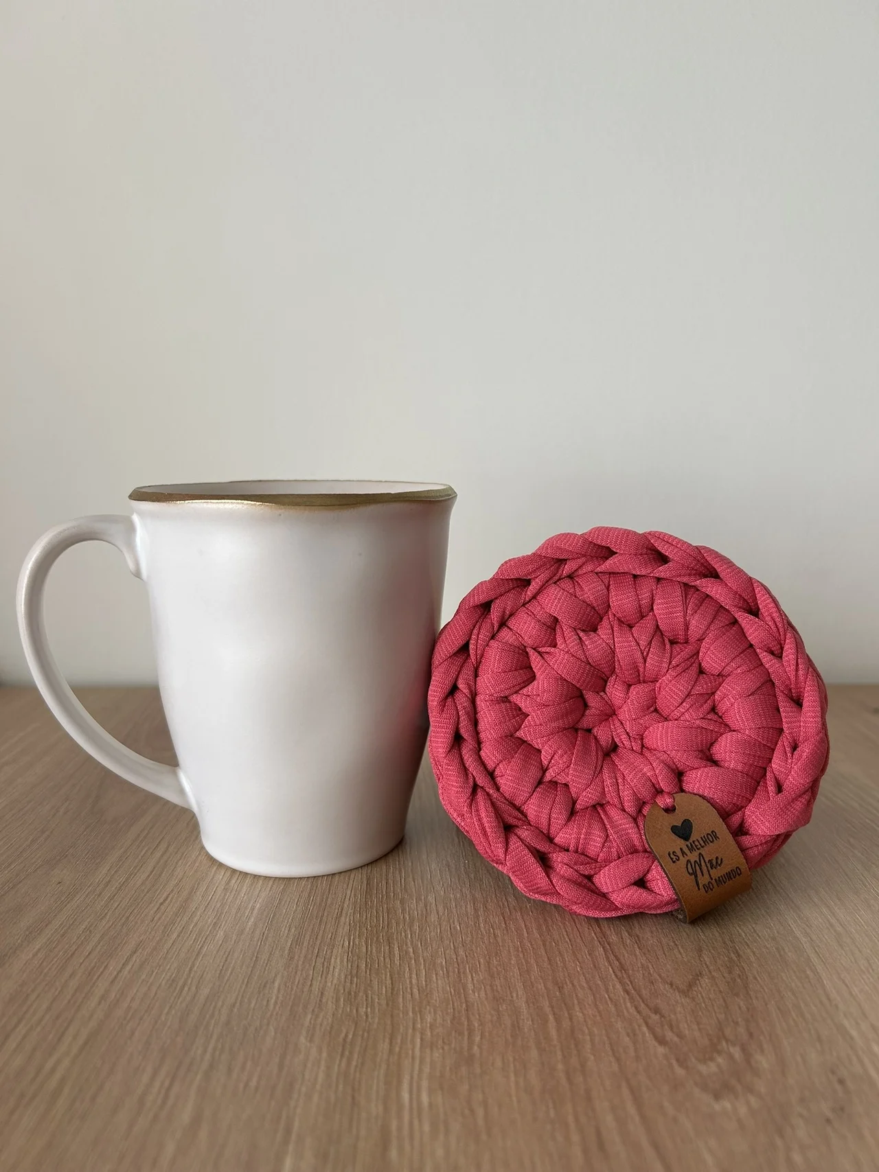 Caneca branca com borda dourada ao lado de suporte trançado rosa com etiqueta em pele numa mesa de madeira