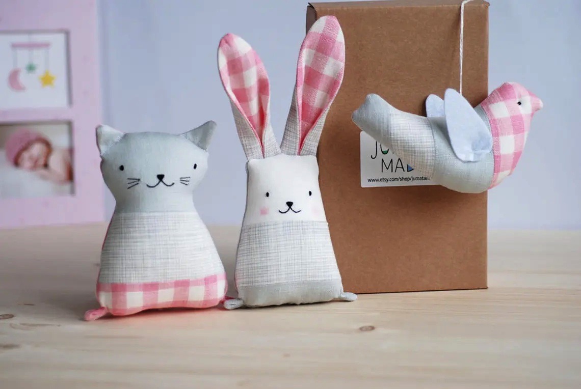 Brinquedos de tecido: gato cinzento, coelho bege com orelhas cor-de-rosa e pássaro cor-de-rosa com asas cinzentas junto a caixa de papel
