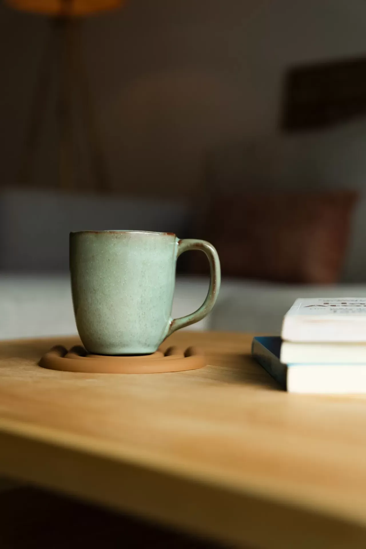 Caneca verde em cerâmica sobre suporte de cortiça numa mesa de madeira com livros desfocados ao fundo