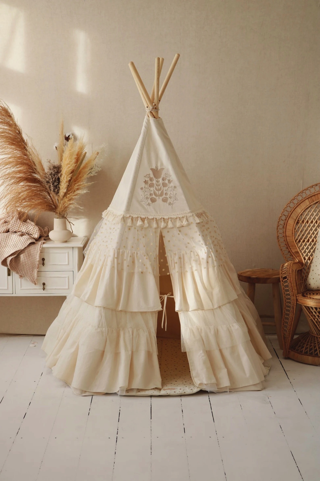 Tenda tipi infantil creme com folhos e padrão floral, em quarto com móveis e cadeira de vime.