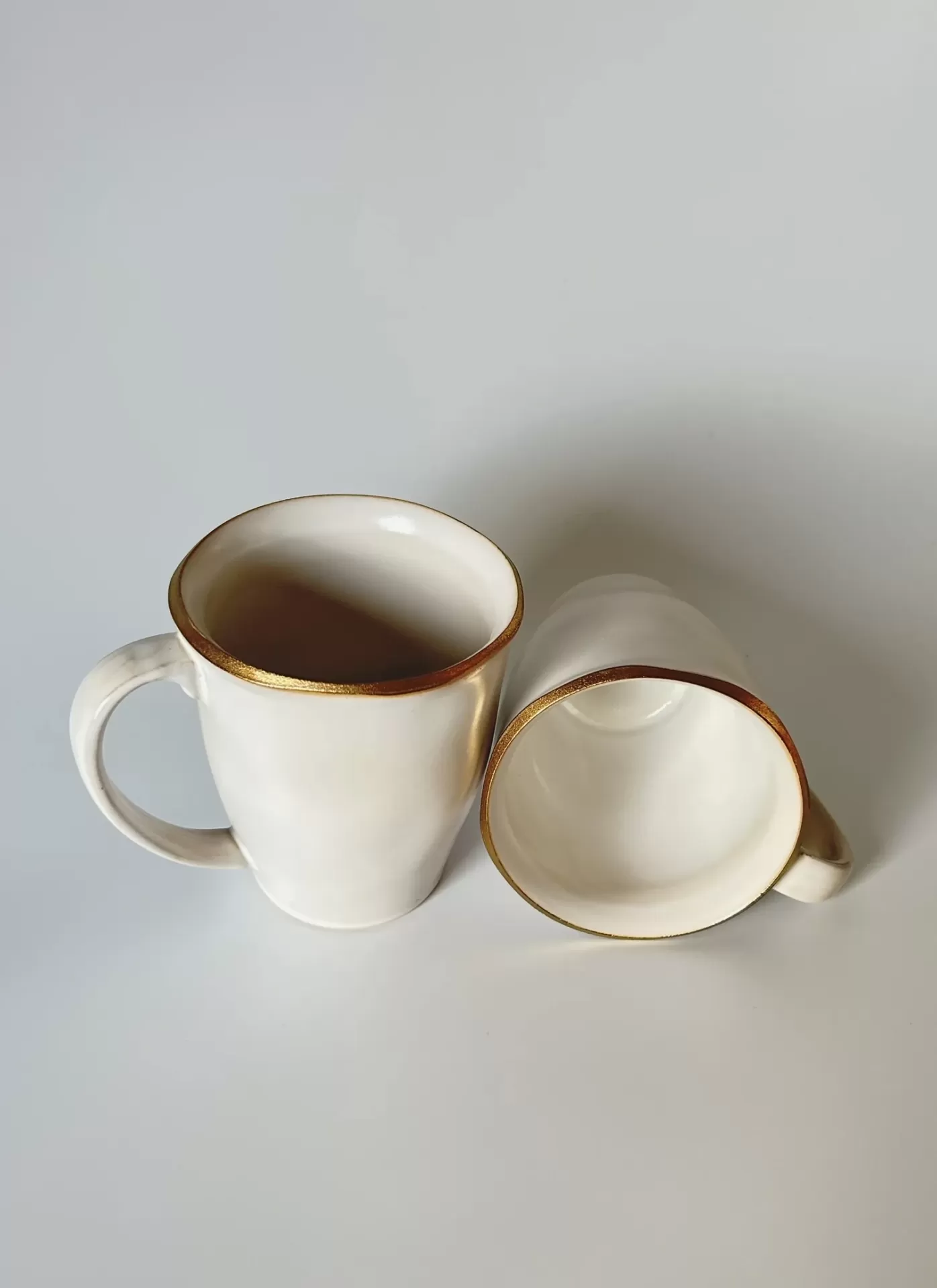 Duas canecas de cerâmica branca com bordas douradas, sobre fundo branco.