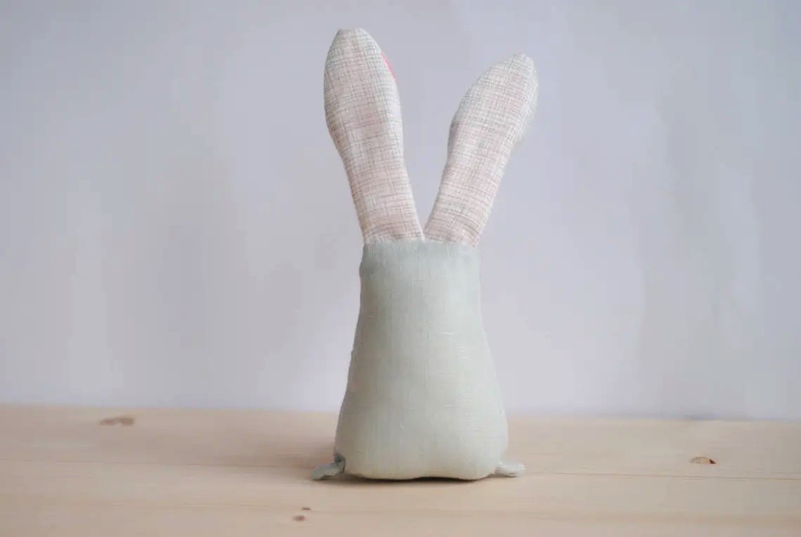 Boneco de peluche em forma de coelho branco com orelhas xadrez rosa e branco em fundo cinzento.