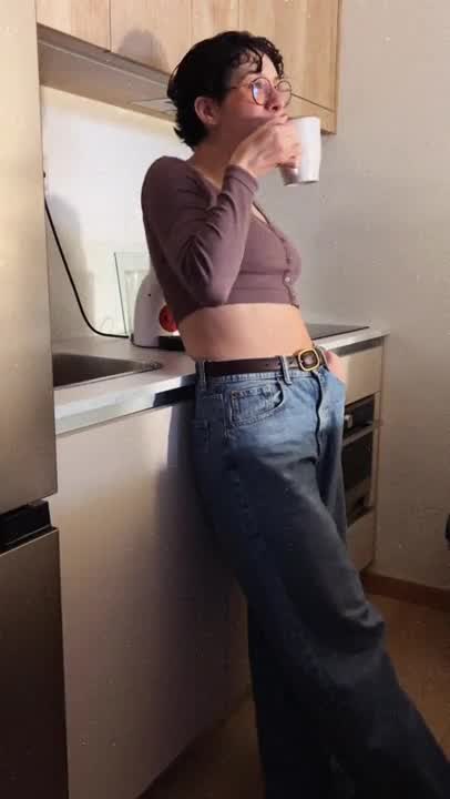 mulher com top castanho e calças azuis larga a beber numa chávena branca numa cozinha moderna