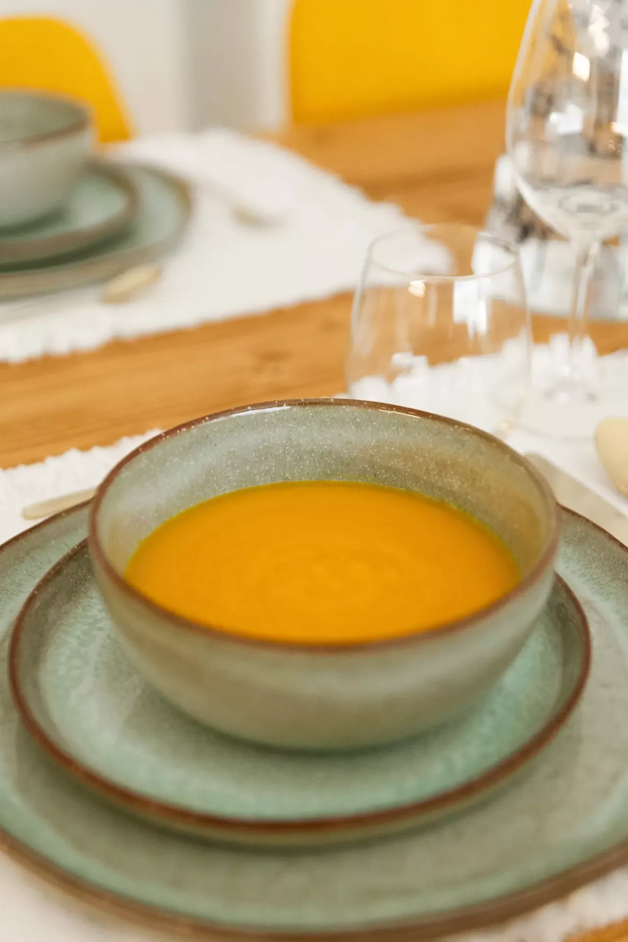 Prato fundo de cerâmica cinza com sopa laranja em mesa com toalha branca e copos de vidro ao fundo