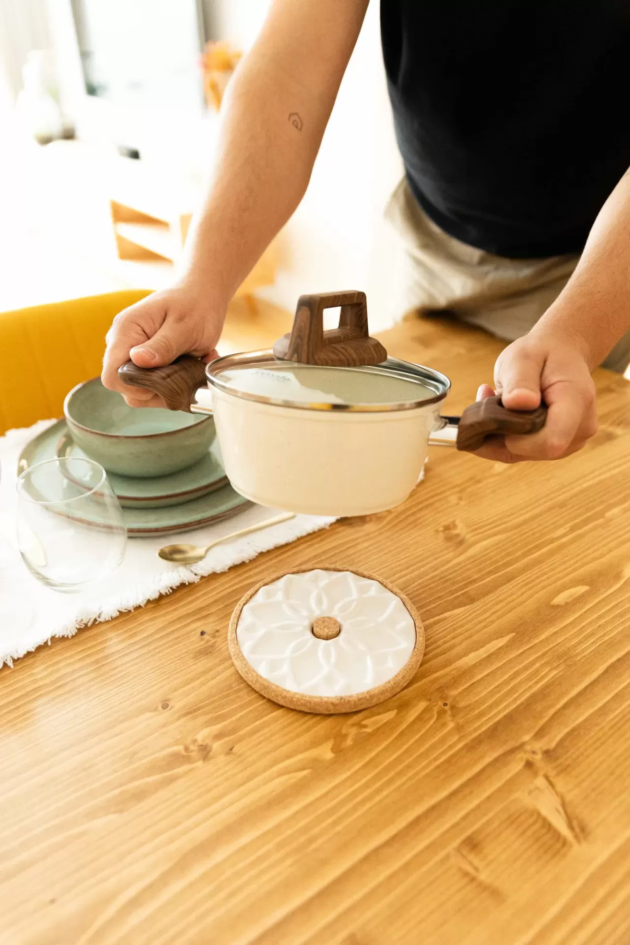 Panela de cozinha branca com tampa de vidro e pegas de madeira em mesa de madeira com louça e descanso para panela.