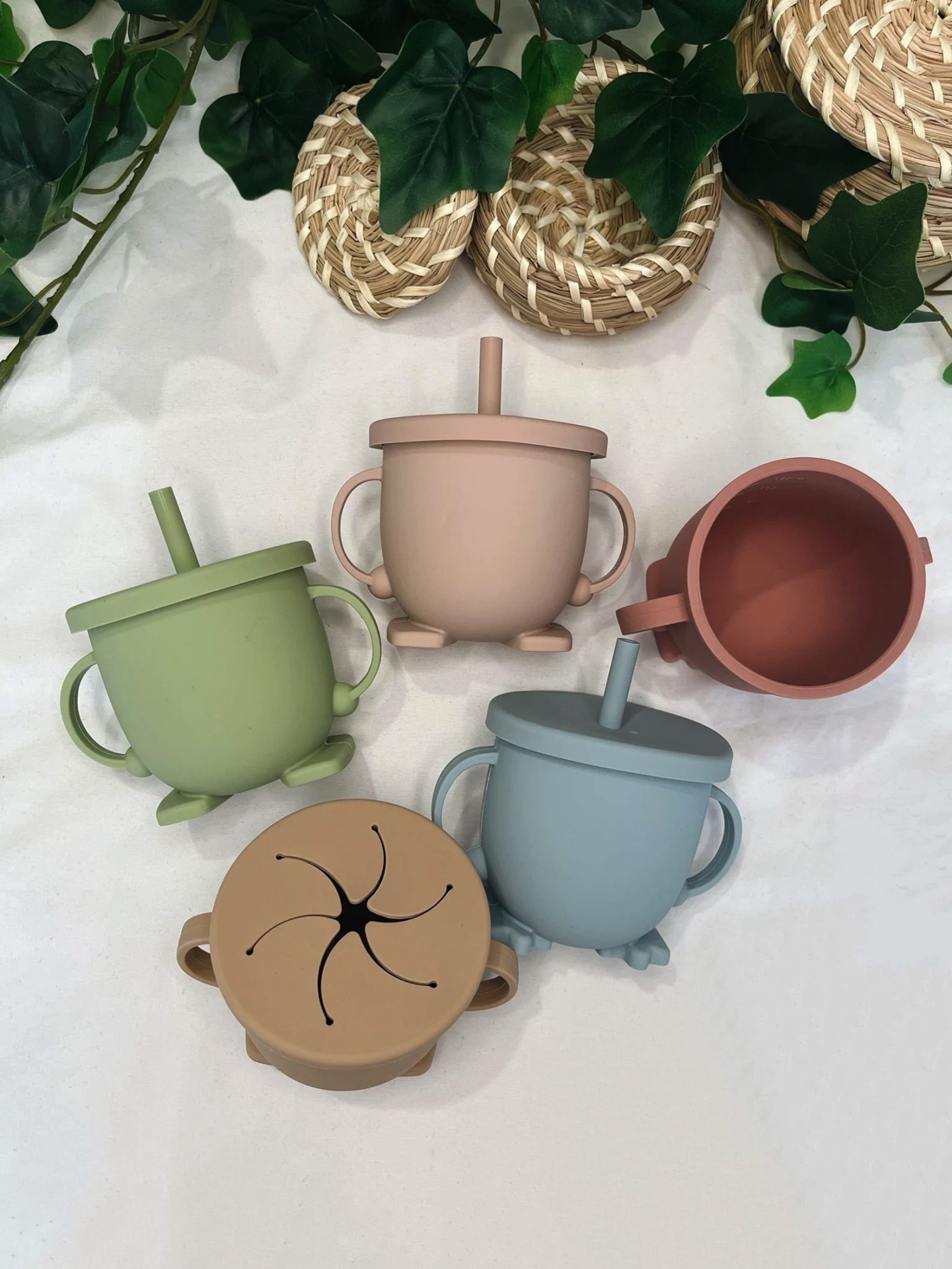 Copos infantis de silicone com canudo e tampas em várias cores, com pegadores duplos, sobre tecido branco e folhas verdes