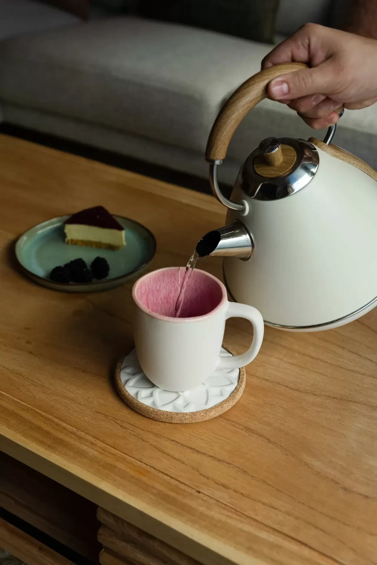 Bule elétrico branco a verter água para chávena branca com interior rosa sobre mesa de madeira com bolo e frutos num prato azul.