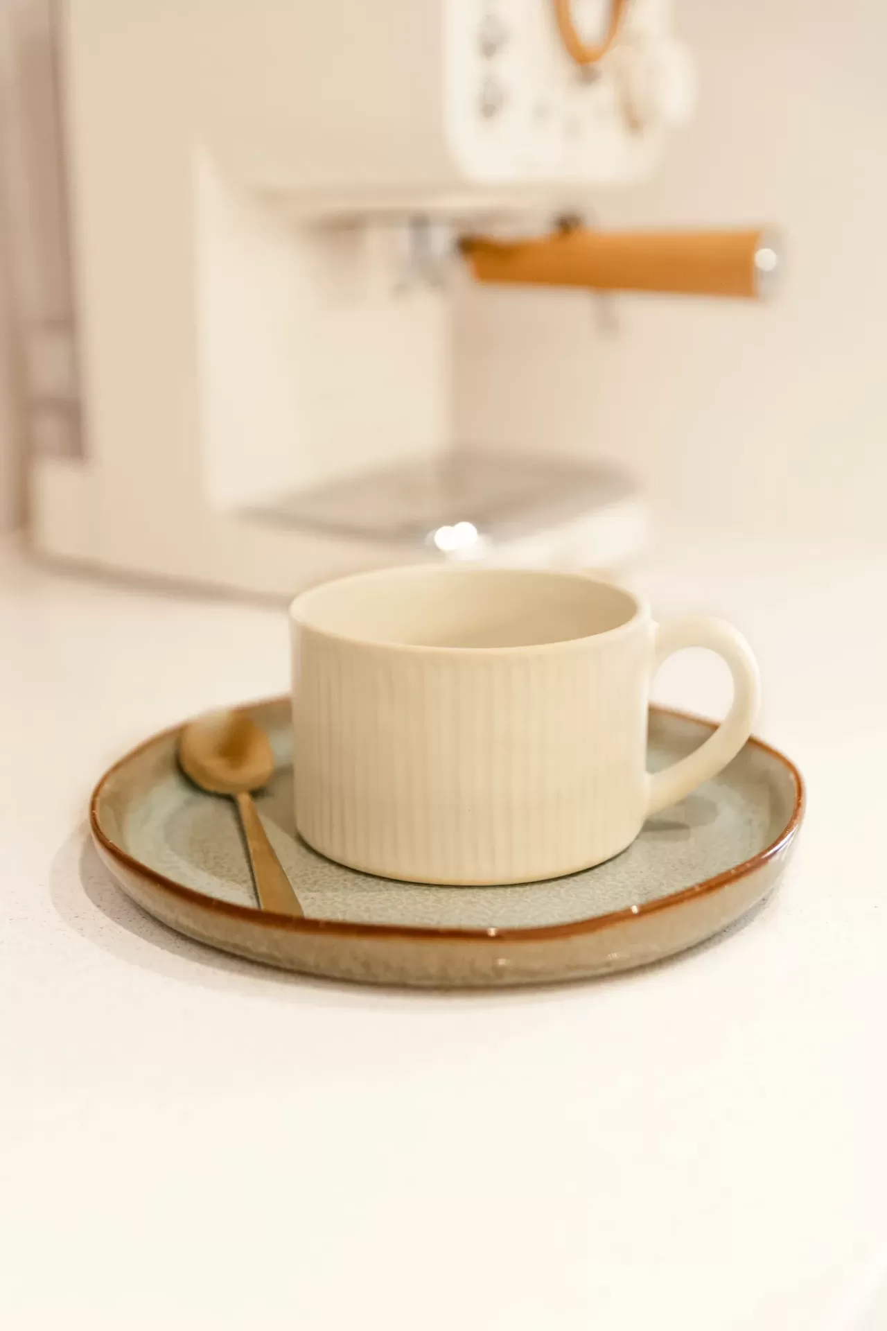 Xícara de cerâmica creme com pires cinza esverdeado e colher dourada ao lado de máquina de café