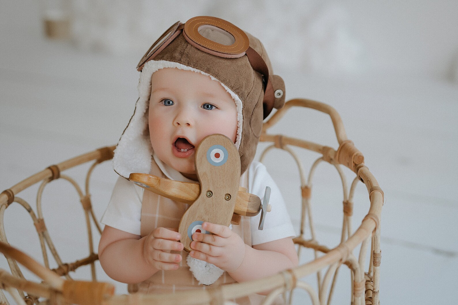Bebé com chapéu aviador castanho segurando brinquedo de avião de madeira dentro de cesto de vime