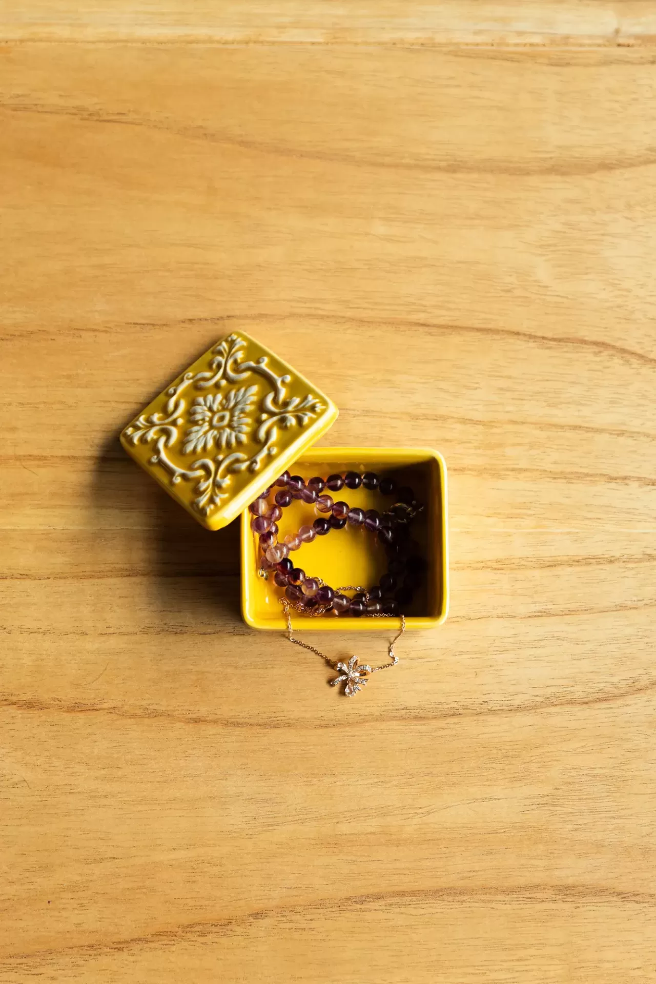 Caixa quadrada amarela decorativa com colares dentro, sobre superfície de madeira clara.