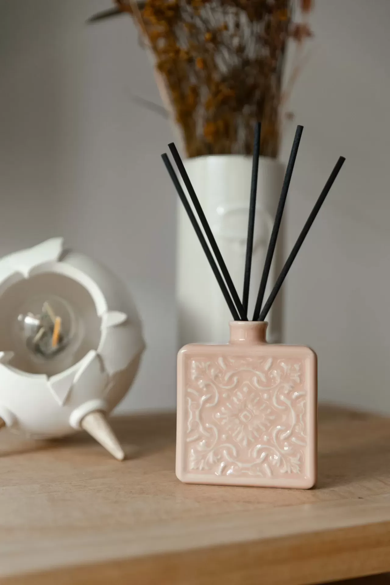 Difusor de ambientador rosa com varetas pretas sobre madeira com fundo de vaso branco e ornamento