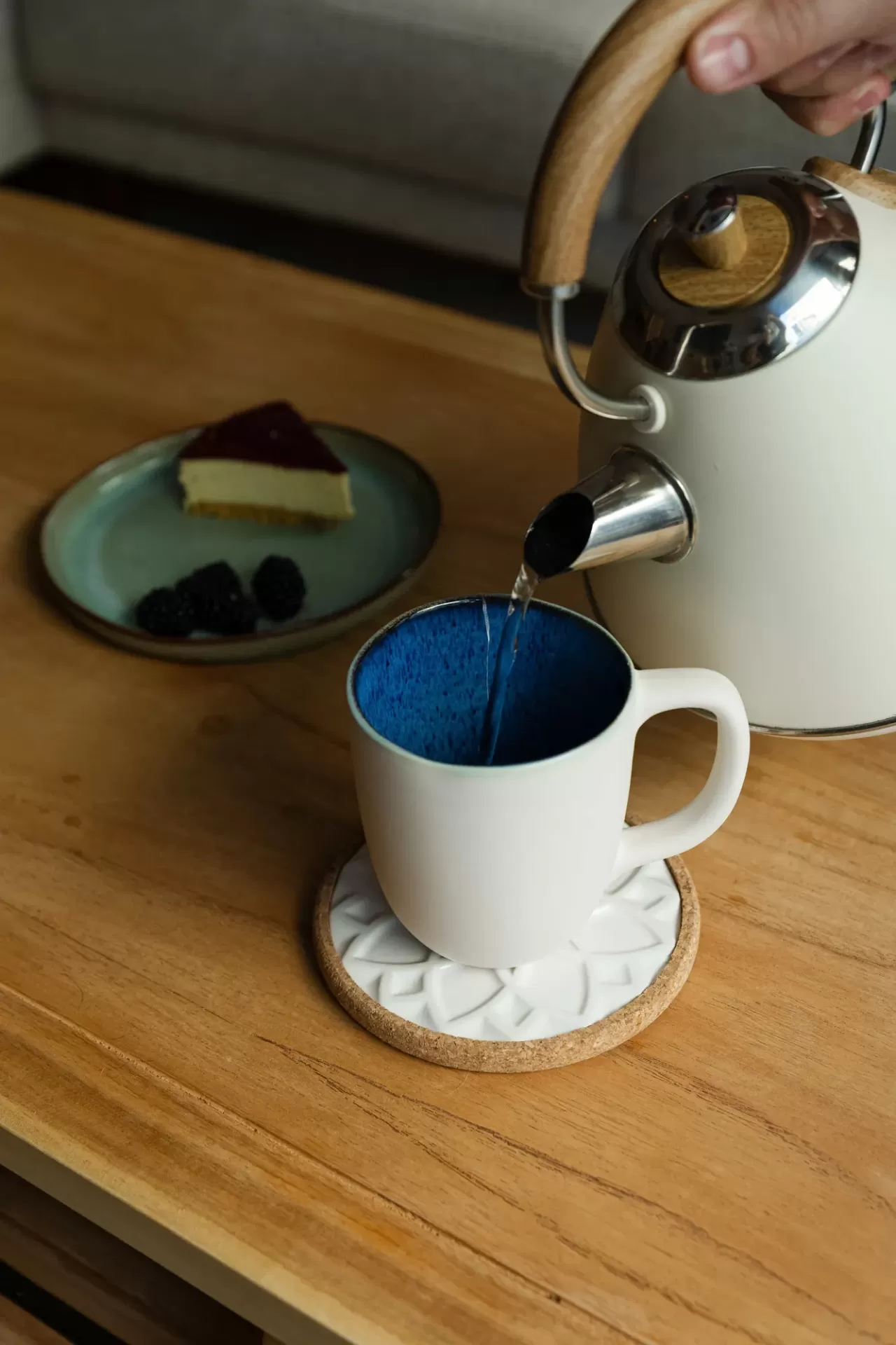Bule elétrico cinzento com detalhes em madeira a verter água para chávena branca com interior azul numa mesa de madeira.