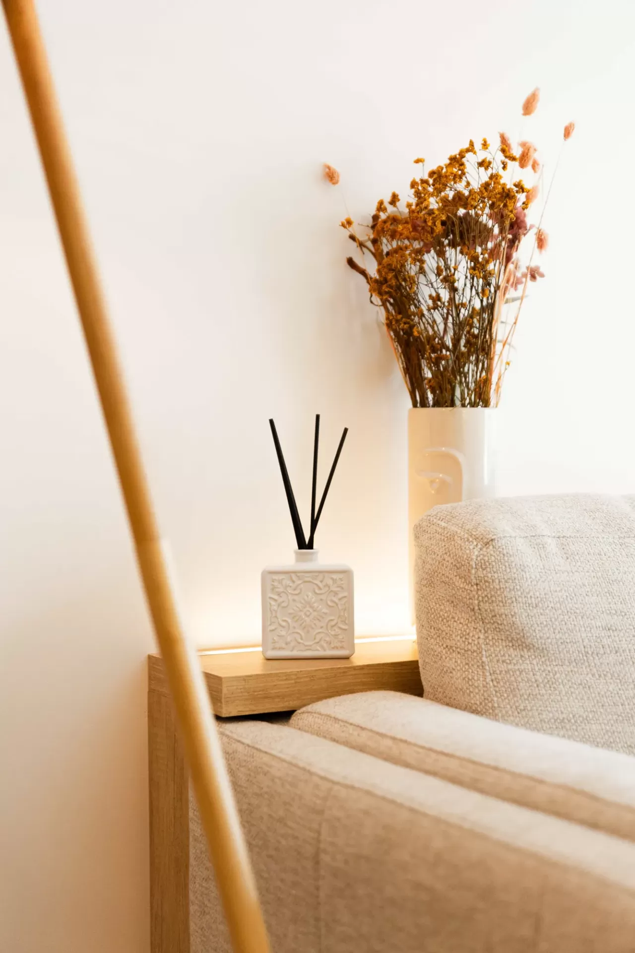 Difusor de fragrância branco com varetas pretas e jarro com flores secas junto a sofá bege texturizado.