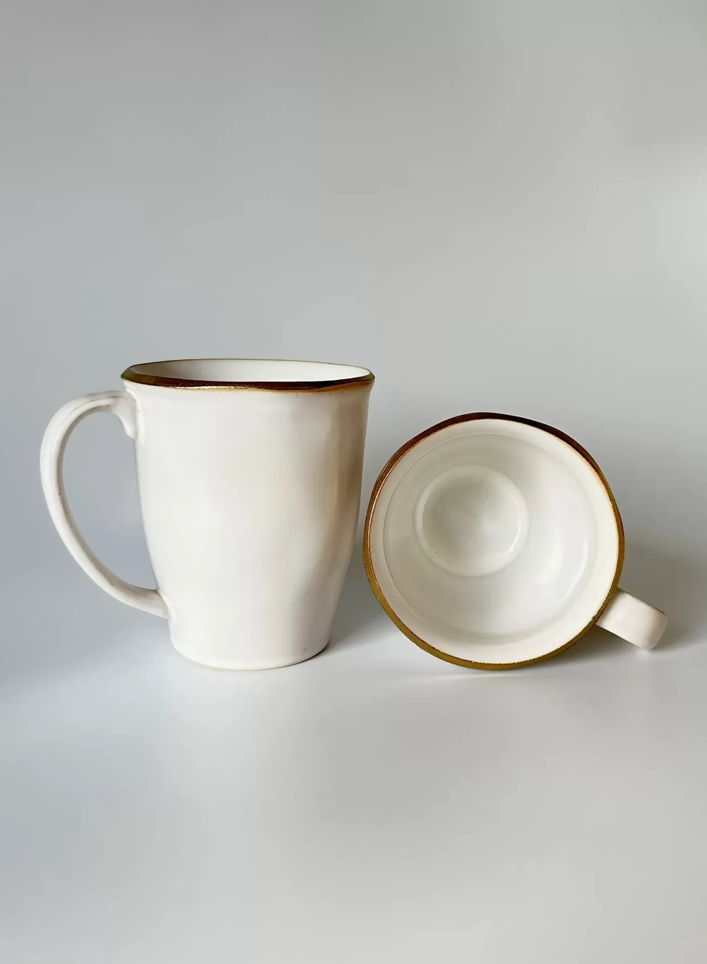 Duas canecas de cerâmica branca com bordas douradas numa superfície branca.